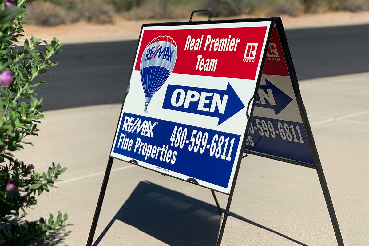 A RE/MAX open house sign featuring the Real Premier Team's branding in red, white, and blue, stands proudly in the driveway of a home. The sign includes the team leader's cell phone number. This image relates to our blog post discussing the effectiveness of open houses in the home selling process