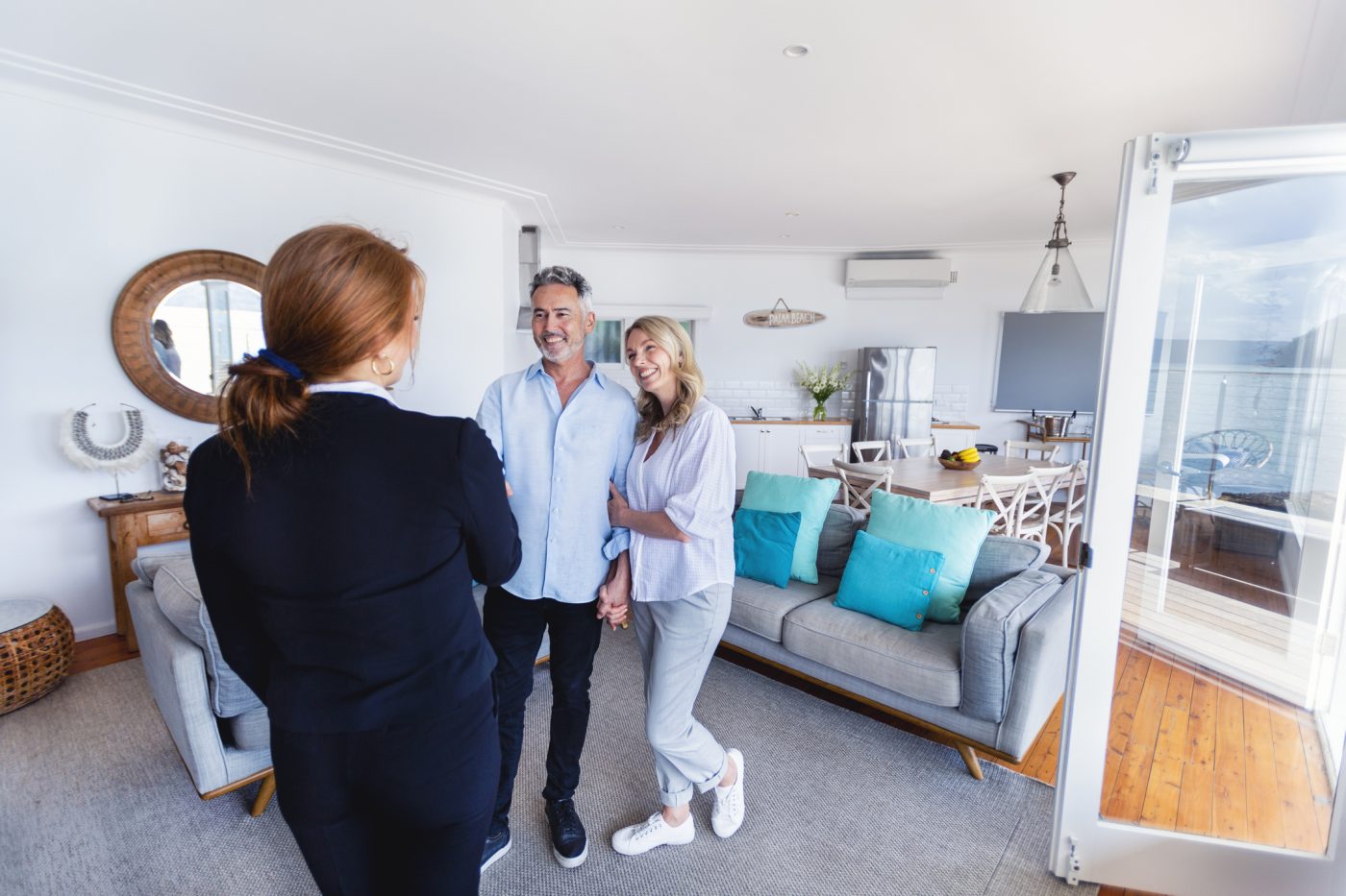 A cheerful red-headed realtor stands in a bright, beautifully updated room, engaging with a mature, delighted couple. The couple embraces as they attentively listen to the realtor, who is sharing valuable information about the housing market. This image captures the essence of concierge customer service, elevating the home buying experience.