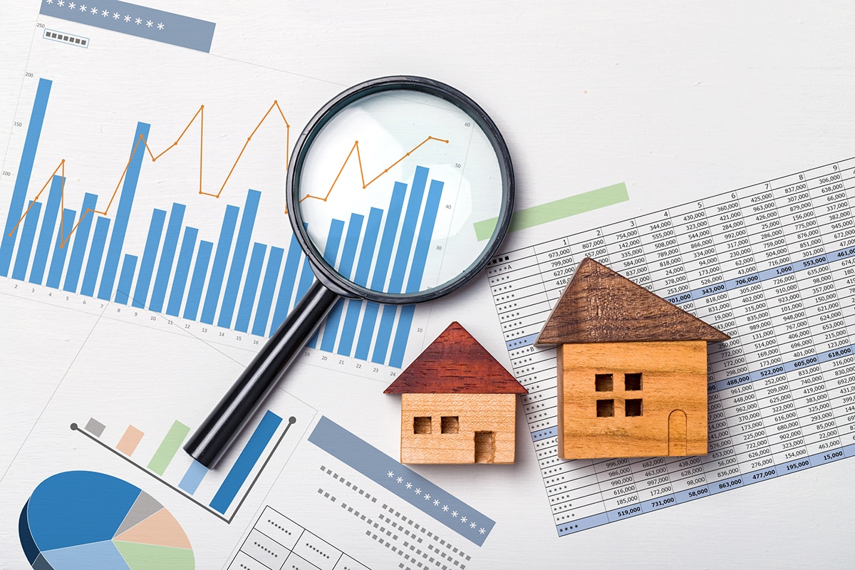 A stack of charts and graphs displaying numbers and figures, illustrating local housing trends. A magnifying glass is positioned on top to signify close examination. In the midst of the spread-out papers, two wooden house-shaped paperweights suggest this is a snapshot of a prospective homebuyer's workspace, taking a momentary break from their research.