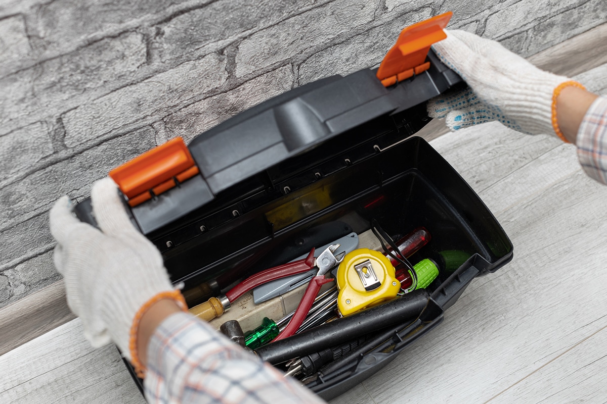 Image of a homeowner opening a basic, single-compartment toolbox, illustrating the essentials of home maintenance. Inside the toolbox, tools such as a tape measure, screwdrivers, a box cutter, a hammer, and pliers are seen lying on top of each other in a somewhat disorganized manner, emphasizing the simplicity and practicality of the toolkit necessary for basic home upkeep