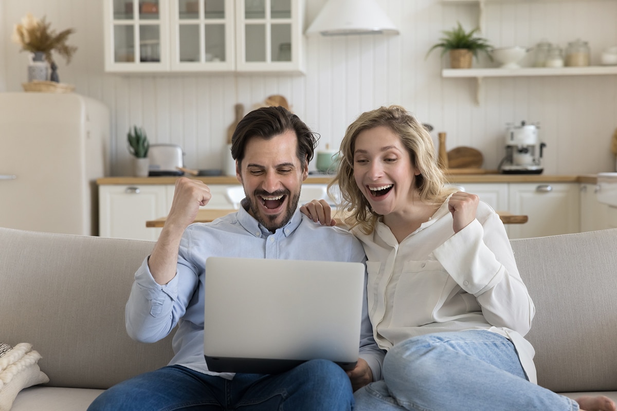 Excited couple sitting closely on a couch, smiling joyfully as they look at a laptop screen. They exhibit visible happiness and satisfaction from their decision to work with Erica from Real Premier Team as their real estate agent. The image captures their elation upon winning the bid for a home they made an offer on, highlighting the success and emotional fulfillment in their real estate journey.
