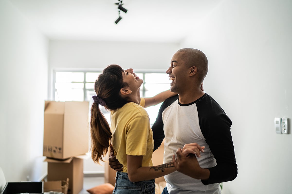 Happy couple embracing indoors, joyfully dancing in an almost empty room with a few unpacked boxes in the background. Their smiles and playful demeanor reflect excitement and contentment, symbolizing a new beginning in their home.