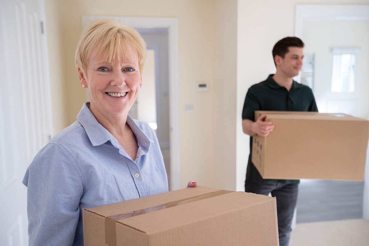 A smiling older woman holding a cardboard box in the foreground with a man carrying a box in the background inside a bright, empty room, symbolizing the start of a downsizing journey.