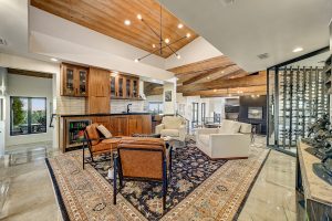 Spacious living room at 6432 E El Sendero Rd featuring wood-beamed ceilings, custom wet bar, modern glass-enclosed wine display, and polished flooring.